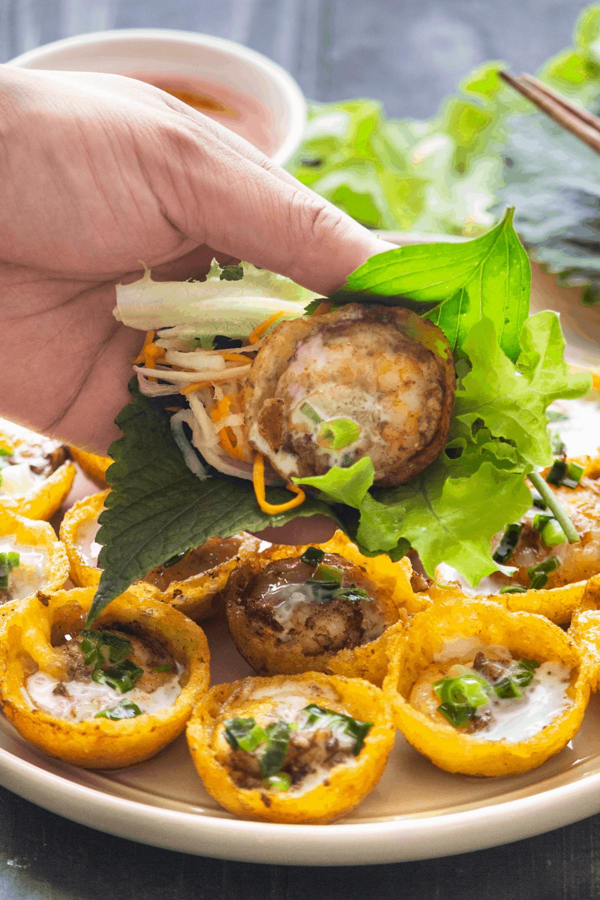 Bánh Khọt on a plate with a hand holding one up wrapped with salad.