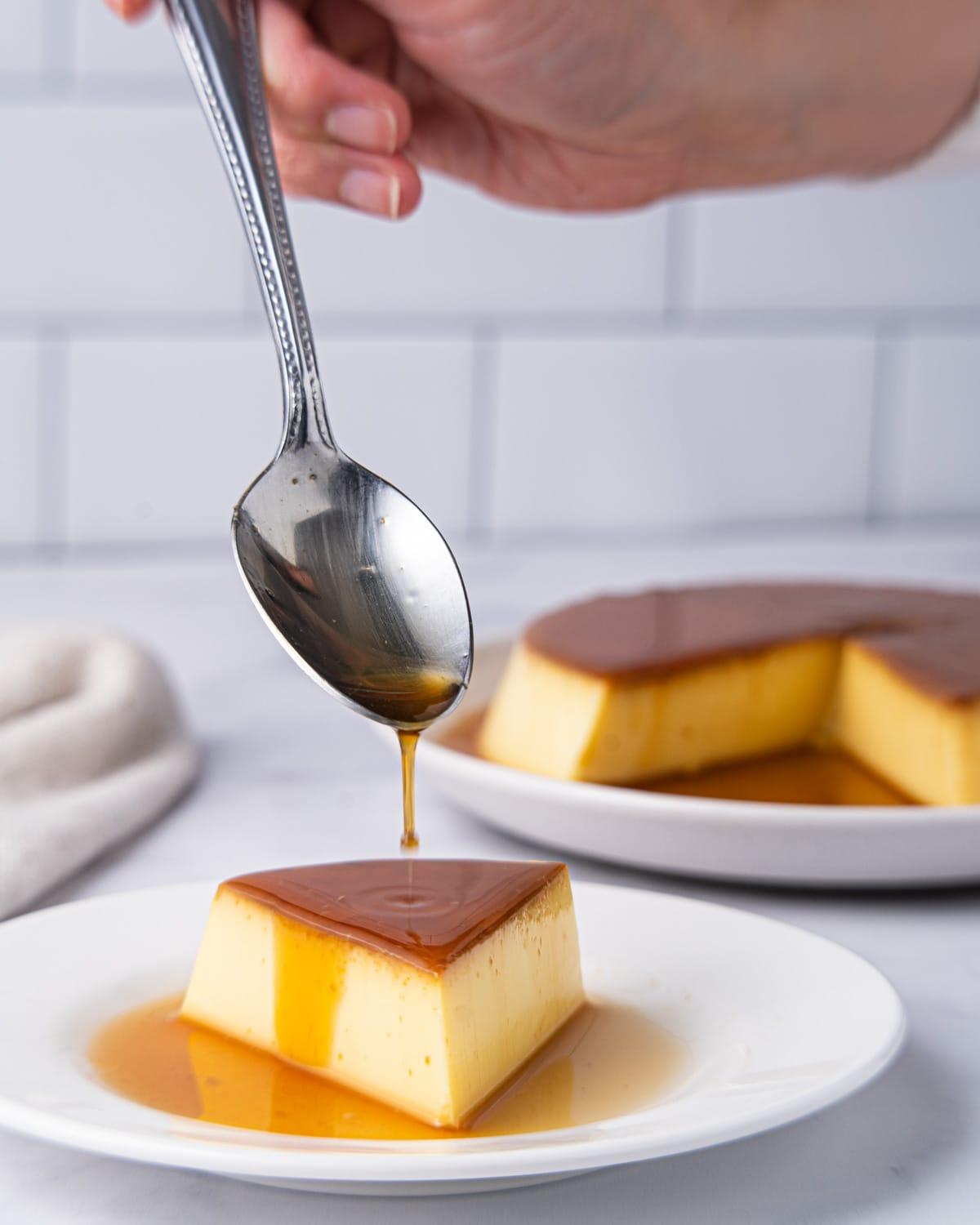 Slice of flan on a plate with spoon drizzling caramel on top.