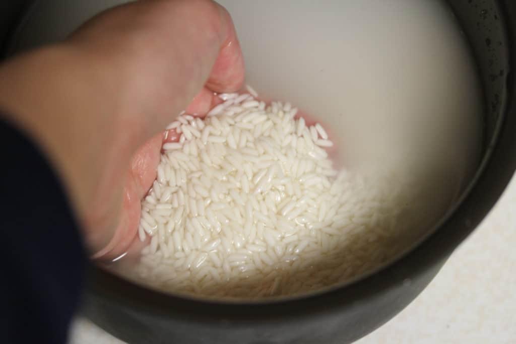 A hand is seen scooping raw rice out of murky water inside of a rice cooker container.