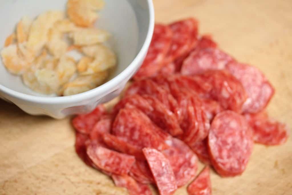 A blue bowl is seen in the upper left hand corner filled with water and dried shrimp. The bowl is on top of a wooden cutting board. Next to the bowl is a pile of thinly sliced chinese sausage
